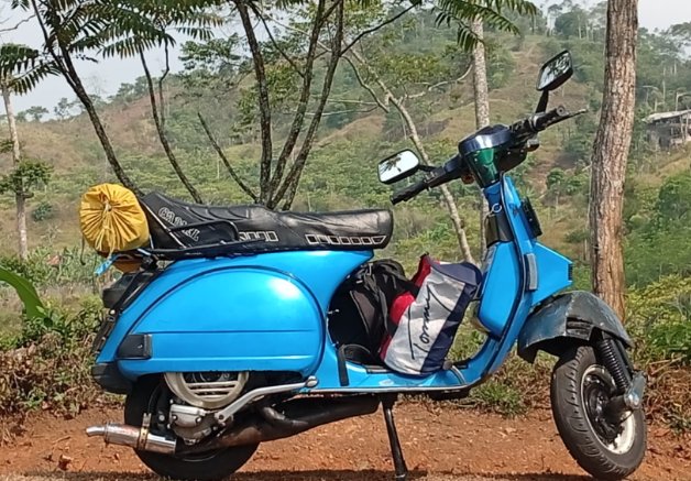 Vespa klasik biru dengan tas dan barang bawaan touring terparkir di jalan tanah perbukitan